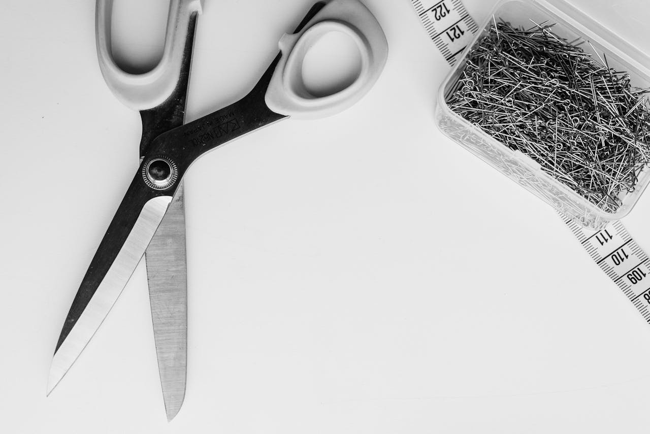 Monochrome image of scissors, measuring tape, and pins on white background.